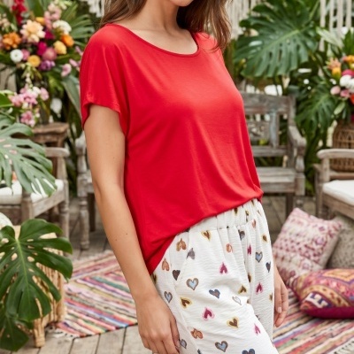 Mulher com blusa vermelha e calças brancas com corações coloridos em ambiente decorado com plantas e almofadas.