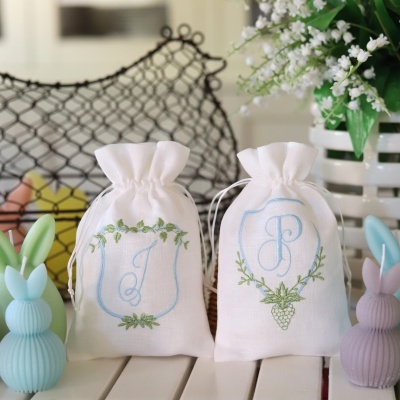 Sacolas de tecido branco bordadas com letras J e P e decorações vegetais, ao lado de coelhos decorativos e planta verde.