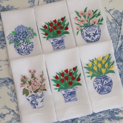 Seis toalhas brancas bordadas com flores coloridas em vasos azuis sobre tecido azul e branco