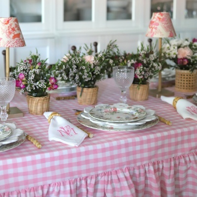 Mesa posta com toalha xadrez rosa e branca, louças florais e vasos com flores em cestos de vime.