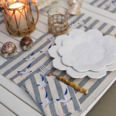Pratos de pétalas brancas empilhados, guardanapo azul e branco com bordados de pássaros, garfo com cabo de bambu, vela em suporte de vidro e ovos decorativos numa mesa branca