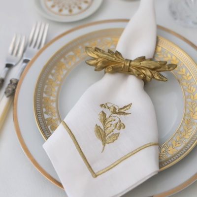 Mesa posta com prato branco e dourado, guardanapo branco com bordado dourado e anel de guardanapo dourado