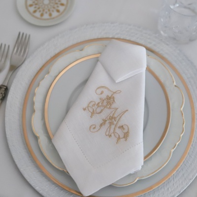 Pratos com bordas douradas, guardanapo branco com monograma AM, talheres e copo num lugar de mesa