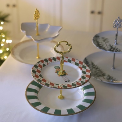Suporte de bolos de dois andares em porcelana com padrões natalícios e pega dourada num fundo branco