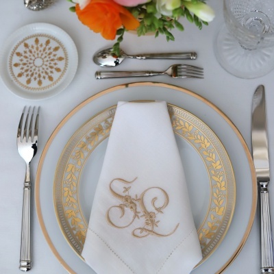 Mesa posta com prato branco e dourado, guardanapo bordado, talheres e flores
