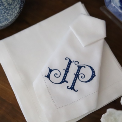 Guardanapo branco com bordado azul das letras 'J P' sobre tecido branco maior em mesa de madeira.