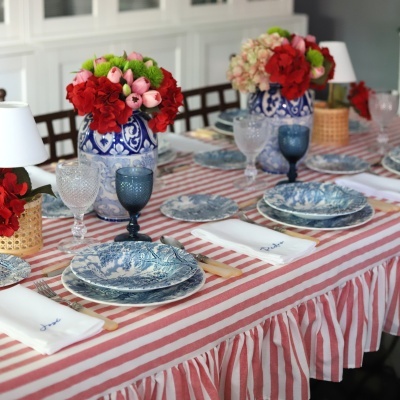 Mesa posta com toalha listrada vermelha e branca, pratos azuis e brancos, copos transparentes e azuis, guardanapos brancos bordados e vasos com flores coloridas