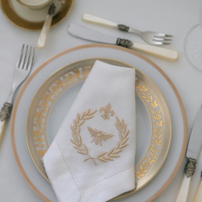Conjunto de jantar com pratos brancos e dourados, guardanapo bordado, talheres com cabos brancos e chávena com pires.