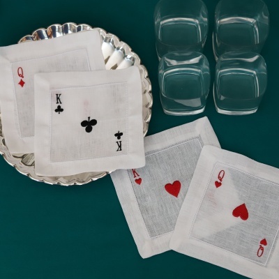 Guardanapos de tecido branco com bordados de cartas de baralho sobre prato prateado e copos quadrados transparentes