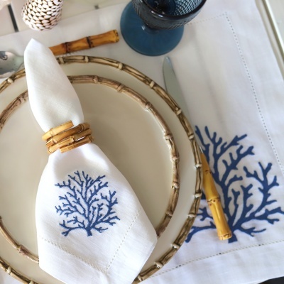 Guardanapo branco com bordado azul de coral e argola em estilo bambu, sobre prato branco com aro bambu, toalhete bordada e talheres com cabo bambu