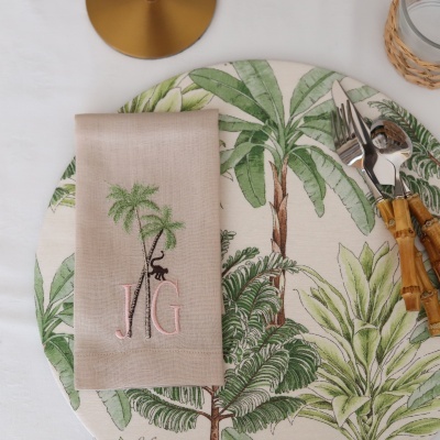 Guardanapo bege bordado com palmeiras e monograma JG sobre prato com padrão de folhas verdes e talheres com cabos em madeira
