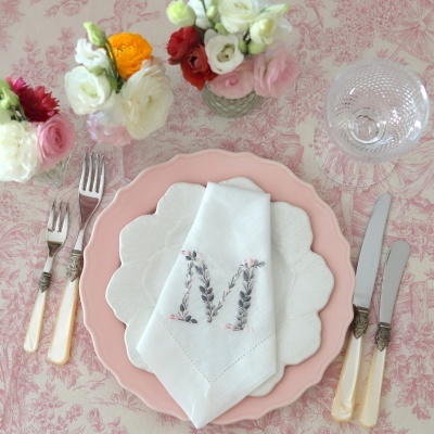 Mesa posta com pratos rosa e branco, guardanapo bordado, talheres com cabos perolados, copo de vidro e flores em vasos sobre toalha floral