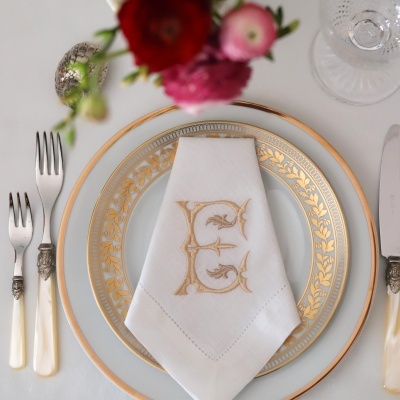 Mesa posta com prato porcelana branca e dourada, guardanapo bordado com letra E, talheres com cabo de madrepérola, copo de vidro e flores coloridas.