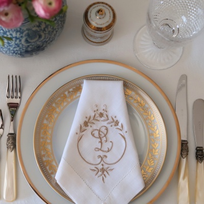 Mesa posta com pratos de porcelana branca e dourada, guardanapo bordado, talheres, copo de vidro e mola decorativa