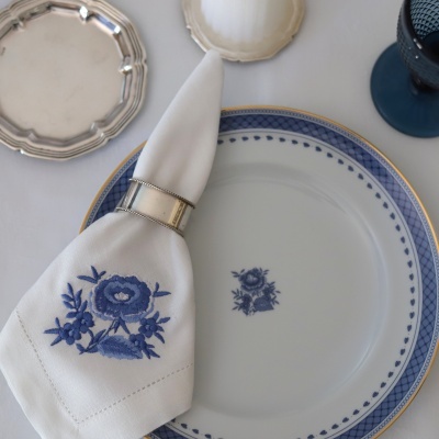 Prato branco com padrão azul e dourado, guardanapo branco com bordado azul de flores e argola prateada, prato pequeno prateado e copo azul