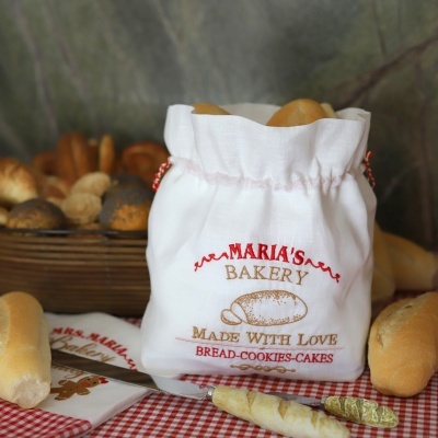 Saco de pano branco bordado com pão, pães ao redor e mesa com toalha xadrez vermelha e branca
