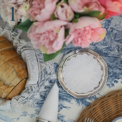 Mesa posta com pão, flor rosa, guardanapo branco, prato prata e toalha azul e branca
