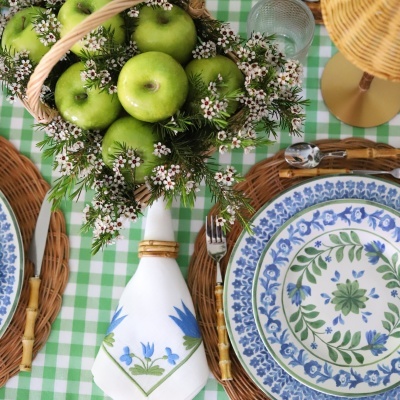 Cesta com maçãs verdes e flores em cima de mesa xadrez verde e branca com pratos e talheres