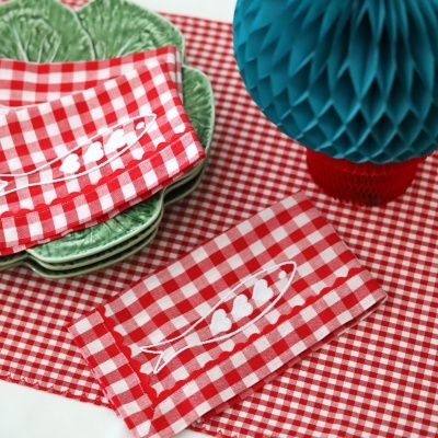 Guardanapos vermelhos e brancos às quadrículas com bordado de peixes, pratos verdes texturados e decoração azul e vermelha sobre toalha vermelha às quadrículas