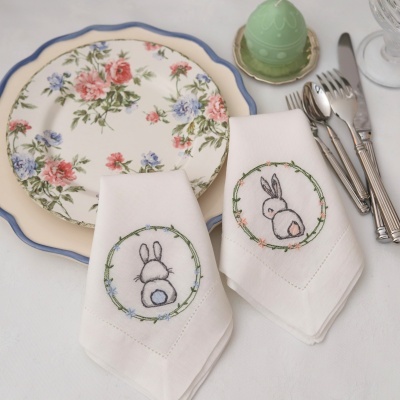 Guardanapos com bordado de coelhos e flores colocados junto a pratos florais e talheres numa mesa.