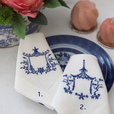 Dois guardanapos brancos bordados a azul sobre prato azul e branco, com vaso de flores e doces cor-de-rosa ao fundo