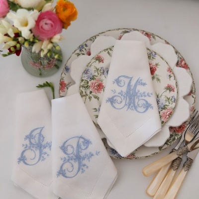 Toalhas brancas bordadas com letras na cor azul sobre pratos florais e talheres com cabo bege