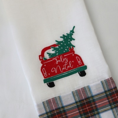 Toalha branca com bordado de carro vermelho e árvore verde, texto Feliz Natal e padrão de quadros coloridos na barra
