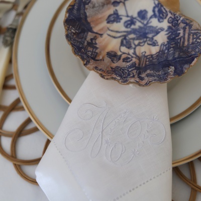 Guardanapo branco com monograma bordado e concha azul escura sobre pratos brancos com borda dourada