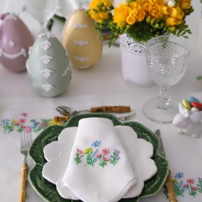 Mesa posta com pratos em verde e branco, guardanapo bordado com flores, talheres com cabo de madeira, copo de vidro, flores amarelas, ovos decorativos e coelho branco com amêndoas.