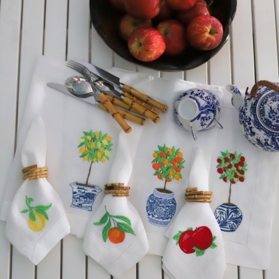 Guardanapos brancos bordados com frutas e vasos azuis numa mesa branca com talheres, chávena, bule e tigela com maçãs