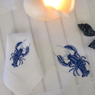 Guardanapos bordados com lagostas azuis, vela em concha acesa, e conchas escuras sobre pano branco com riscas.