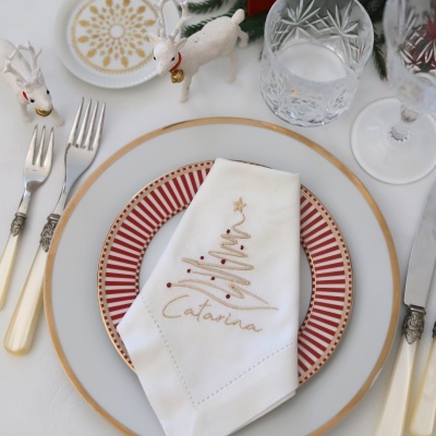 Mesa posta com prato branco com borda dourada, prato pequeno vermelho e dourado, guardanapo branco bordado, talheres creme, dois veados decorativos brancos e copos de vidro lapidado.