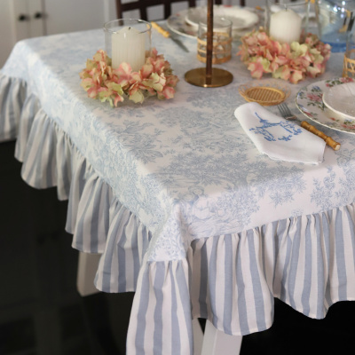 Toalha de mesa azul com padrão floral e saia riscada azul e branca numa mesa posta. Velas com decoração floral artificial.