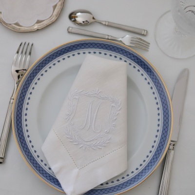 Mesa posta com prato branco com borda azul e dourada, guardanapo branco com monograma, talheres de metal e copo de vidro.