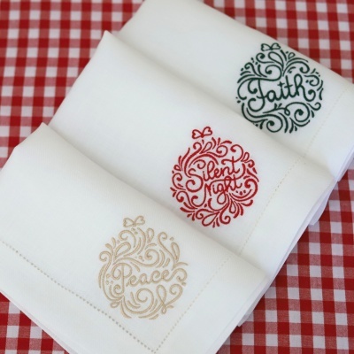 Três tecidos brancos com bordados Faith, Silent Night e Peace, sobre superfície xadrez vermelha e branca.