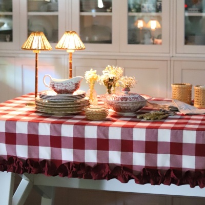 Mesa com toalha de mesa xadrez vermelha e branca, conjunto de loiça branca e vermelha, lâmpadas de mesa e flores em vasos