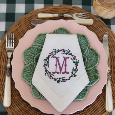 Mesa posta com prato rosa e prato verde em forma de folhas, guardanapo branco bordado à letra M, talheres com cabo branco e fundo de mesa em rattan.