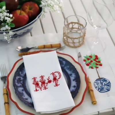 Conjunto de mesa com pratos azuis e creme, guardanapo branco com monograma MP em vermelho, toalha bordada, talheres de bambu e copos transparentes