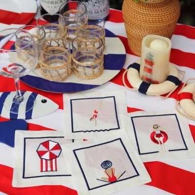 Guardanapos bordados náuticos sobre toalha listrada com copos e decoração náutica.