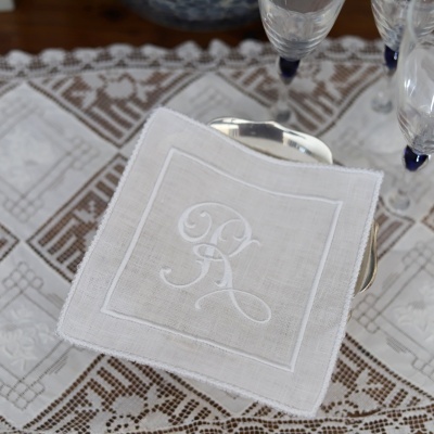 Guardanapo branco bordado com letras colocada sobre prato prateado em toalha branca com padrão castanho.