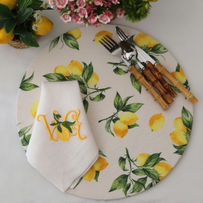 Base de tecido redonda com estampado de limões, guardanapo bordado branco e talheres com cabo de madeira.