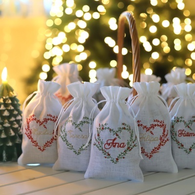 Sacos de tecido branco bordados com nomes e corações, sobre mesa branca com cesto e luzes desfocadas no fundo.