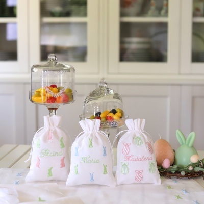 Três saquinhos de tecido branco bordados com nomes e padrões coloridos, suportes de vidro com frutas artificiais e velas decorativas na mesa.