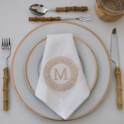 Prato branco com borda dourada, guardanapo com bordado, talheres com cabo de bambu e copo em cesto de rattan.