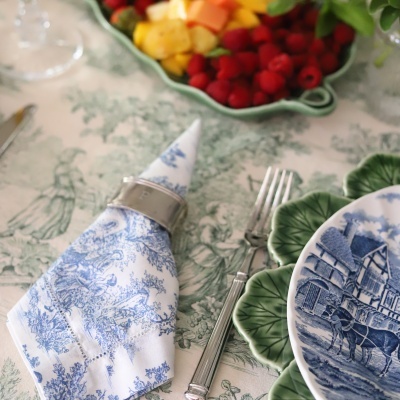 Guardanapo branco com padrão azul num anel prateado junto a pratos verdes e metálicos em mesa decorada com frutas ao fundo