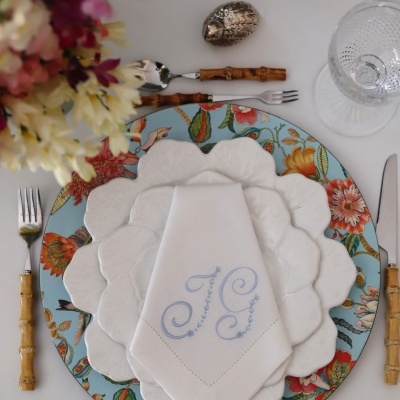 Mesa posta com conjunto de louça branca com borda ondulada sobre prato floral azul, talheres com cabos imitando bambu, guardanapo branco com bordado azul e copo transparente texturizado.