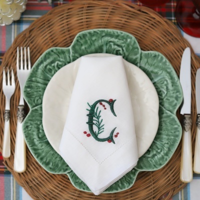 Conjunto de jantar com prato branco, prato verde com textura de folhas, guardanapo branco bordado e talheres com cabo creme, sobre base de rattan e toalha xadrez.