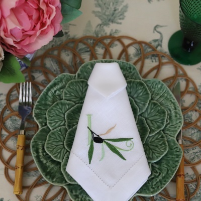 Pratos verdes em forma de folha com guardanapo branco bordado e talheres ao lado sobre base de rattan