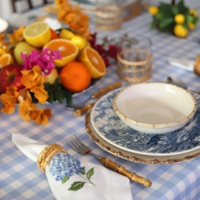 Mesa posta com pratos azul e branco, guardanapo bordado, argolas de bambu, talheres e centro de mesa com frutas e flores.