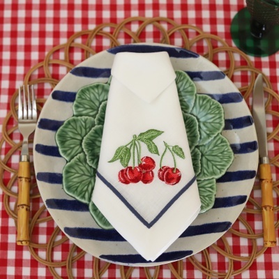 Mesa posta com prato cerâmico listrado azul e branco, prato com padrão folhas verdes, guardanapo branco bordado com cerejas, talheres com cabo de bambu e toalha xadrez vermelha e branca